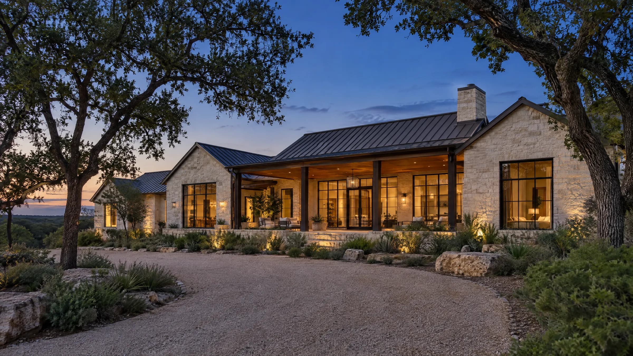 Custom Hill Country home in cream limestone and dark standing-seam metal, windows glowing at dusk under live oaks
