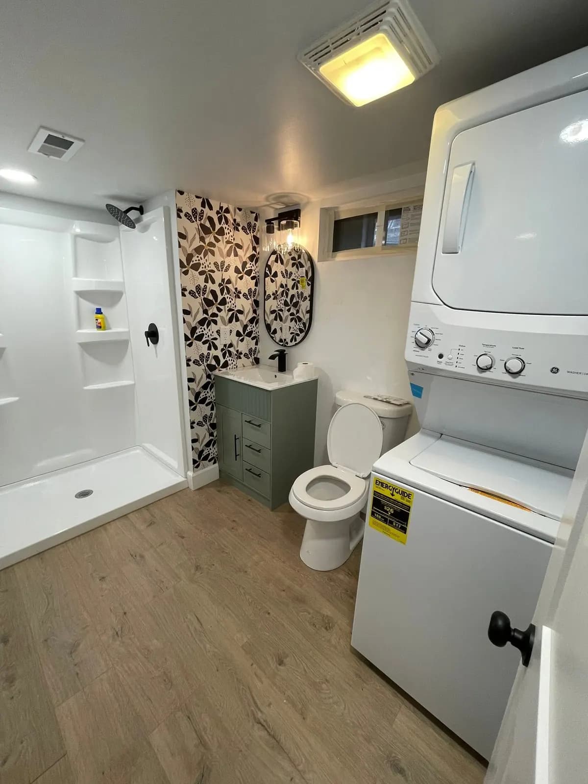 After: the finished bath with a walk-in shower, floral wallpaper wall, sage vanity, and stacked washer-dryer