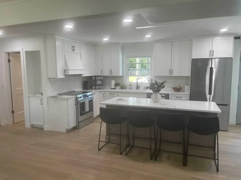 After: the finished kitchen with white shaker cabinets, a quartz island with black stools, and the same bay window now flooding the room